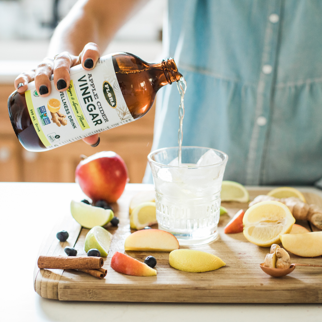 Apple cider vinegar wellness drink with lemon and ginger being poured into cup of ice surrounded by fresh fruit and herbs: apples, lemons, cinnamon, elderberries, and ginger